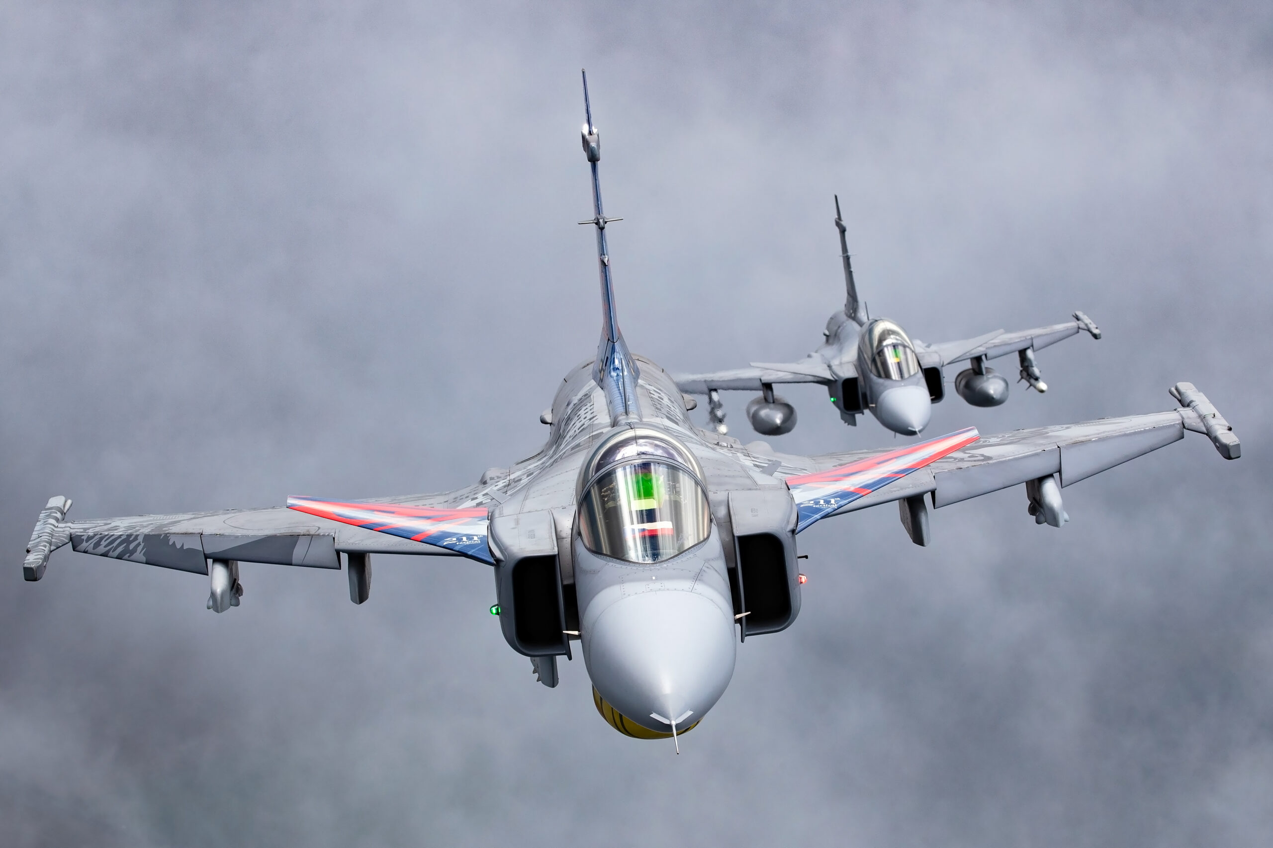 Two Czech Air Force Gripens head-on against grey cloud, the lead jet wearing red, white and blue anniversary stripes on its wings
