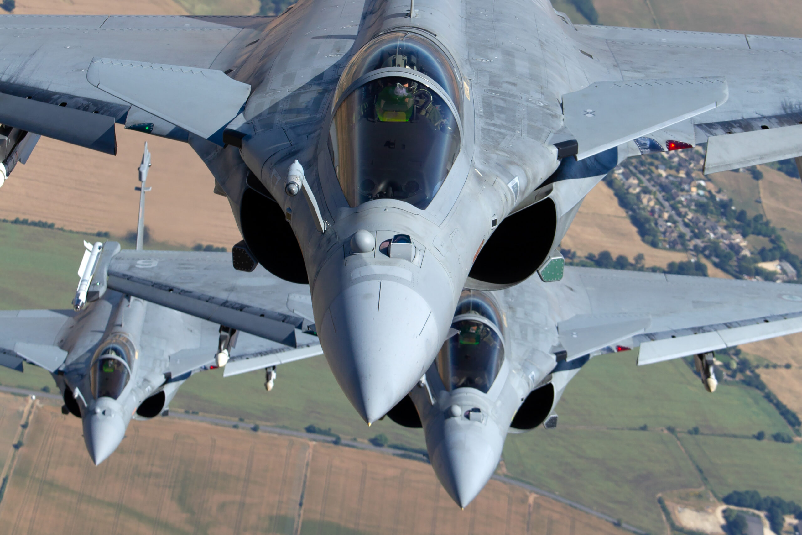 Three French Dassault Rafales stacked in close head-on formation over farmland, pilots visible in the cockpits