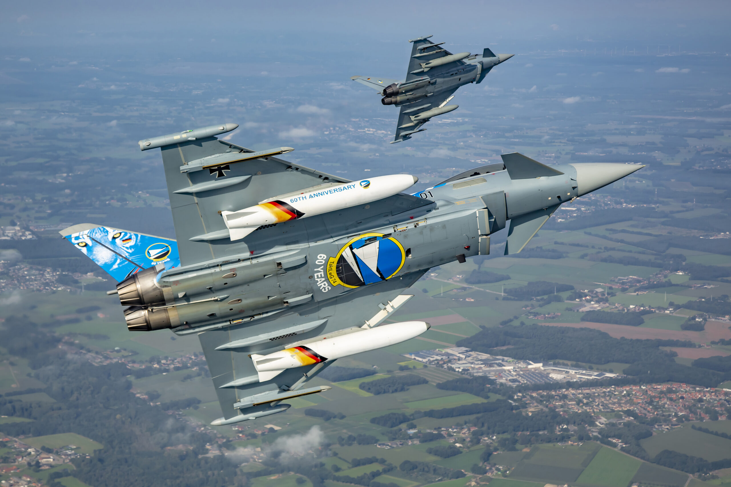 Luftwaffe Typhoon with a blue tiger-faced 60th anniversary TaktLwG 74 tail banking away underside-on, white anniversary fuel tanks visible, with a second Typhoon behind