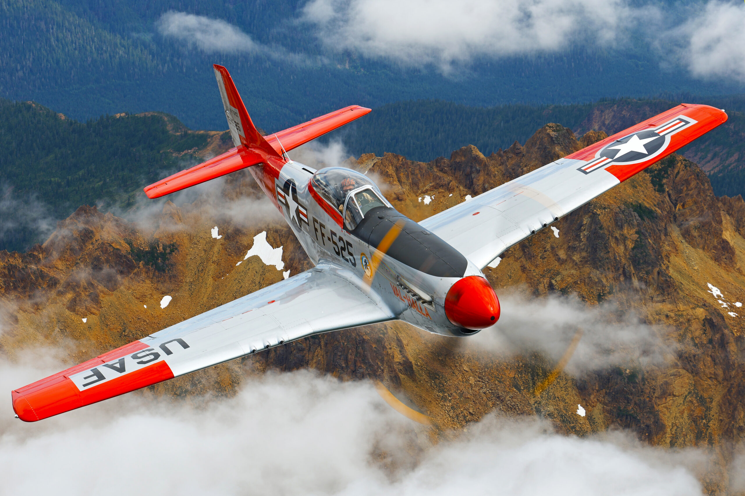 Polished-metal P-51D Mustang Val-Halla with red tail flying over rugged mountains and cloud