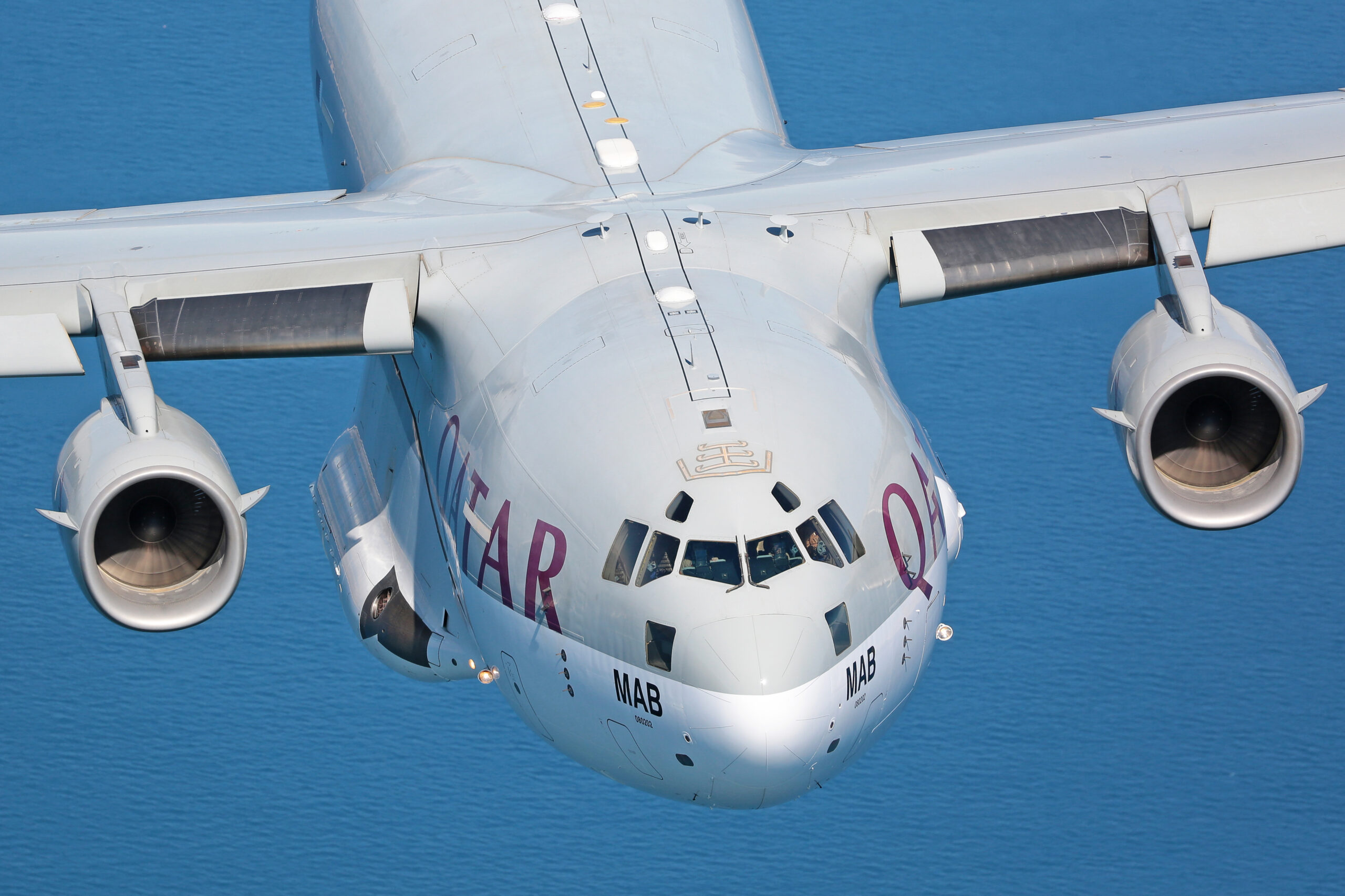 Tight head-on close-up of a Qatar Emiri Air Force C-17A over the sea, QATAR titles and MAB codes visible, crew in the cockpit