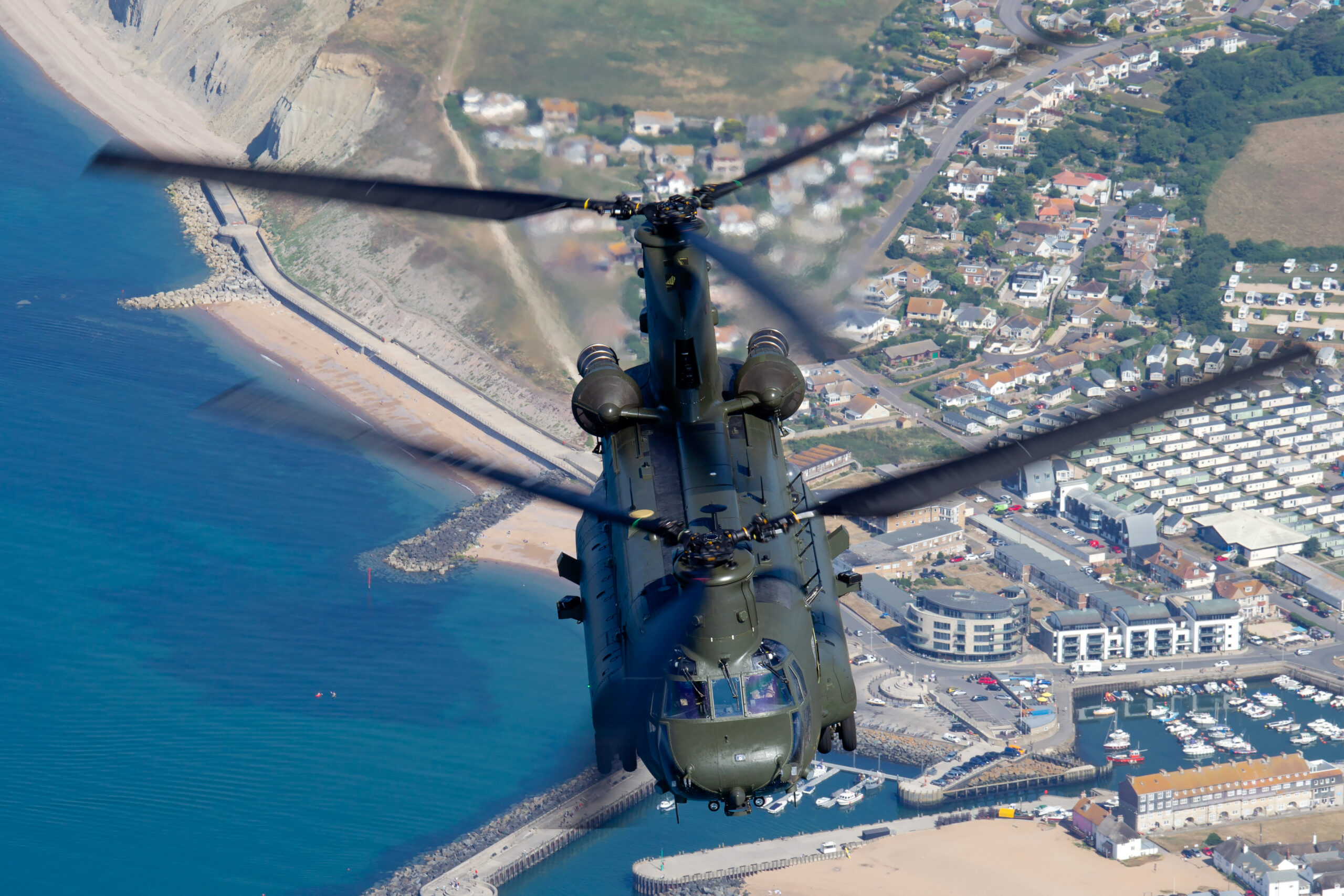 RAF Chinook HC6A head-on over a harbour town and beach, both tandem rotors blurred into full arcs