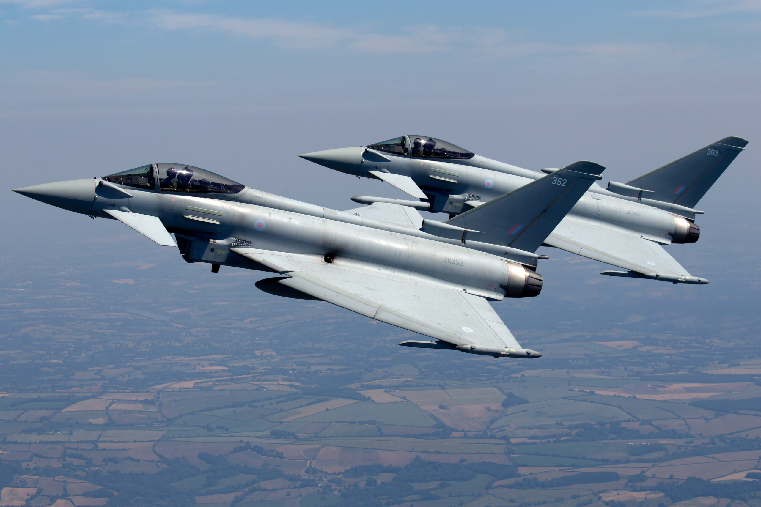 Two RAF Typhoon FGR4s, ZK352 and ZK363, in close echelon formation over hazy patchwork countryside, both pilots visible