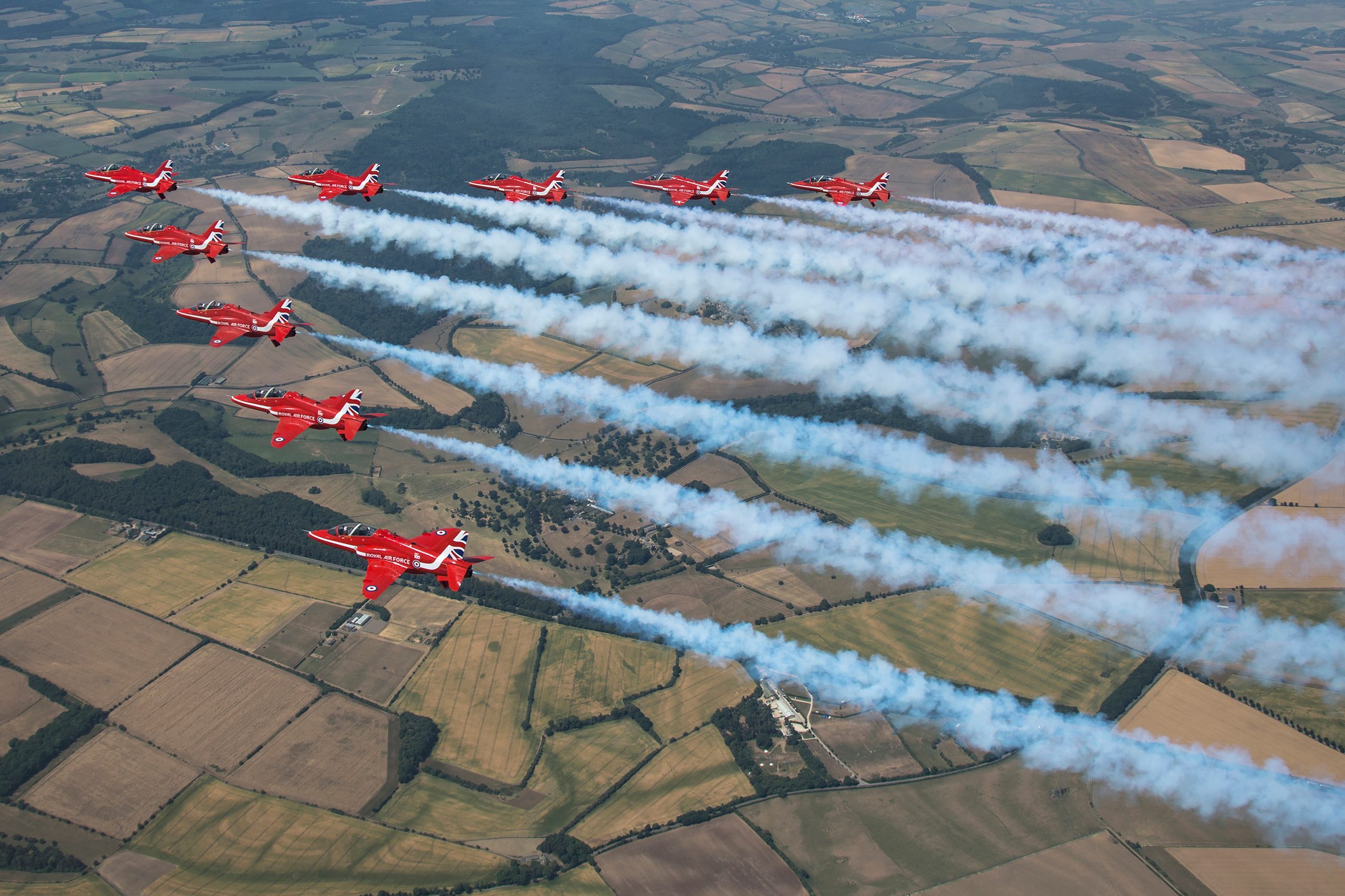 Red Arrows Hawk T1 jets in line abreast trailing coloured smoke over fields