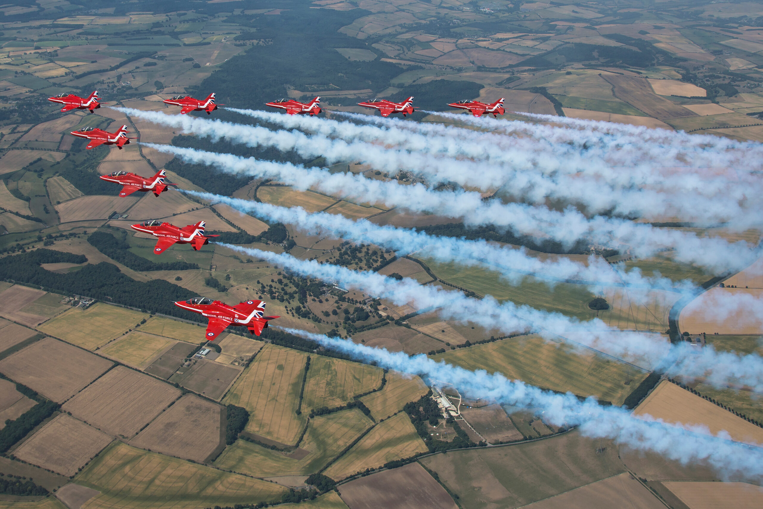 Nine Red Arrows Hawk T1s in formation trailing long white smoke over summer fields, seen from above