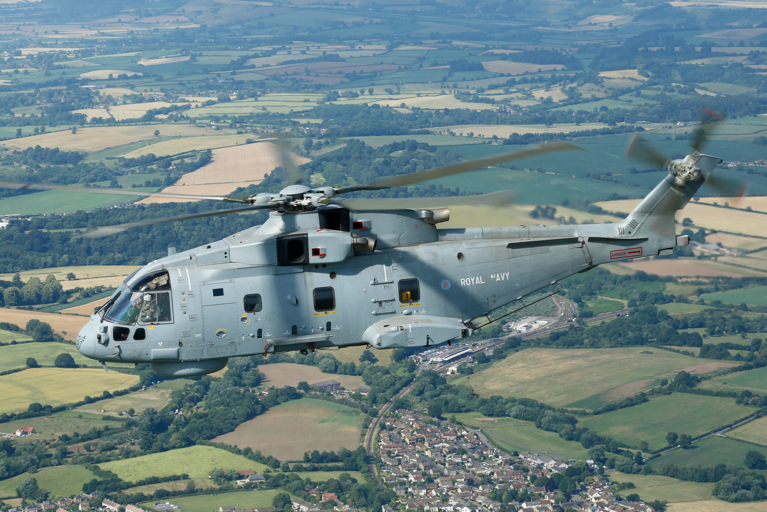 Royal Navy Merlin HM2 helicopter flying over a patchwork of green and gold fields, rotors blurred