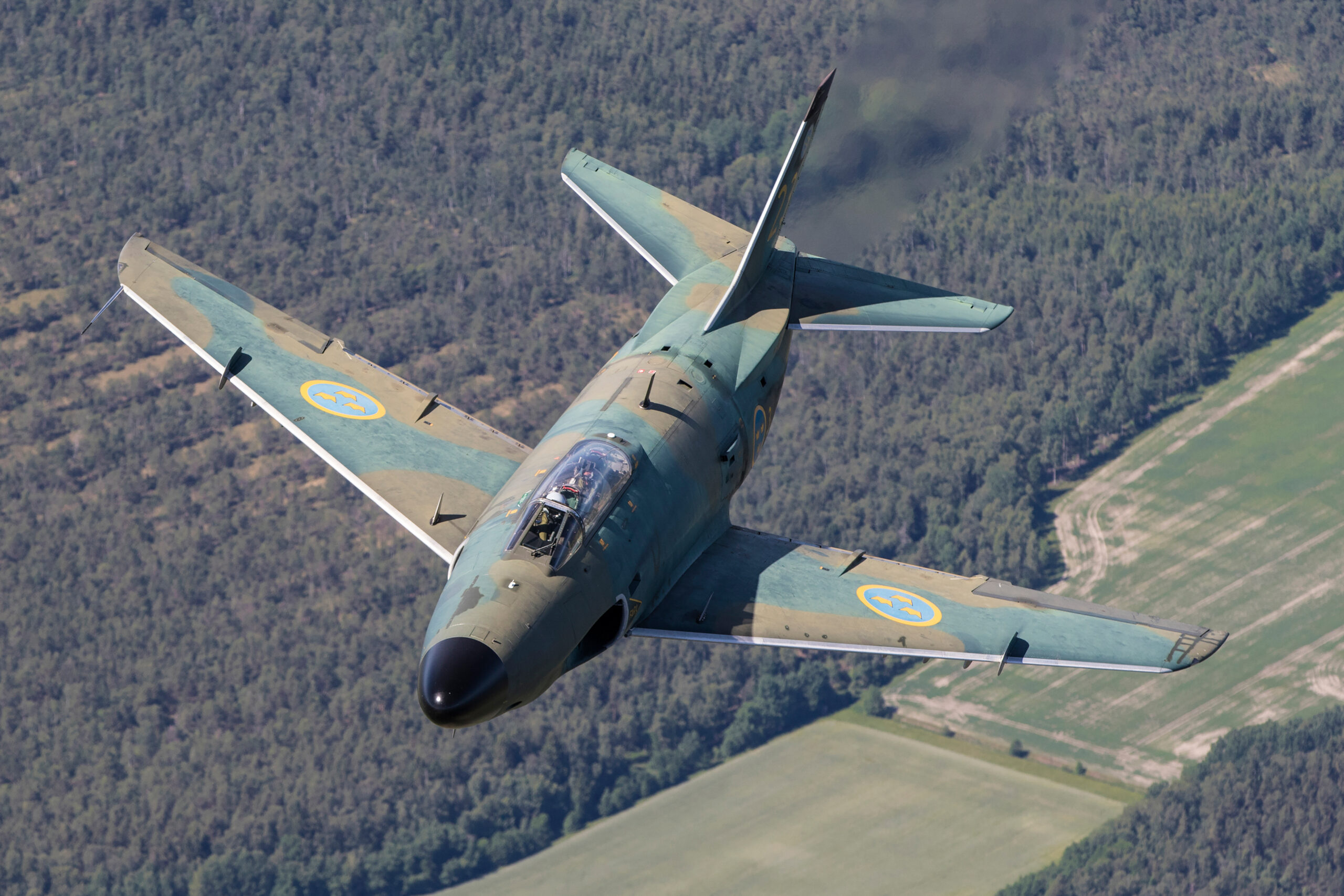 Camouflaged Saab 32 Lansen banking over forest and fields with dark exhaust smoke trailing, pilot visible in the open-visor cockpit