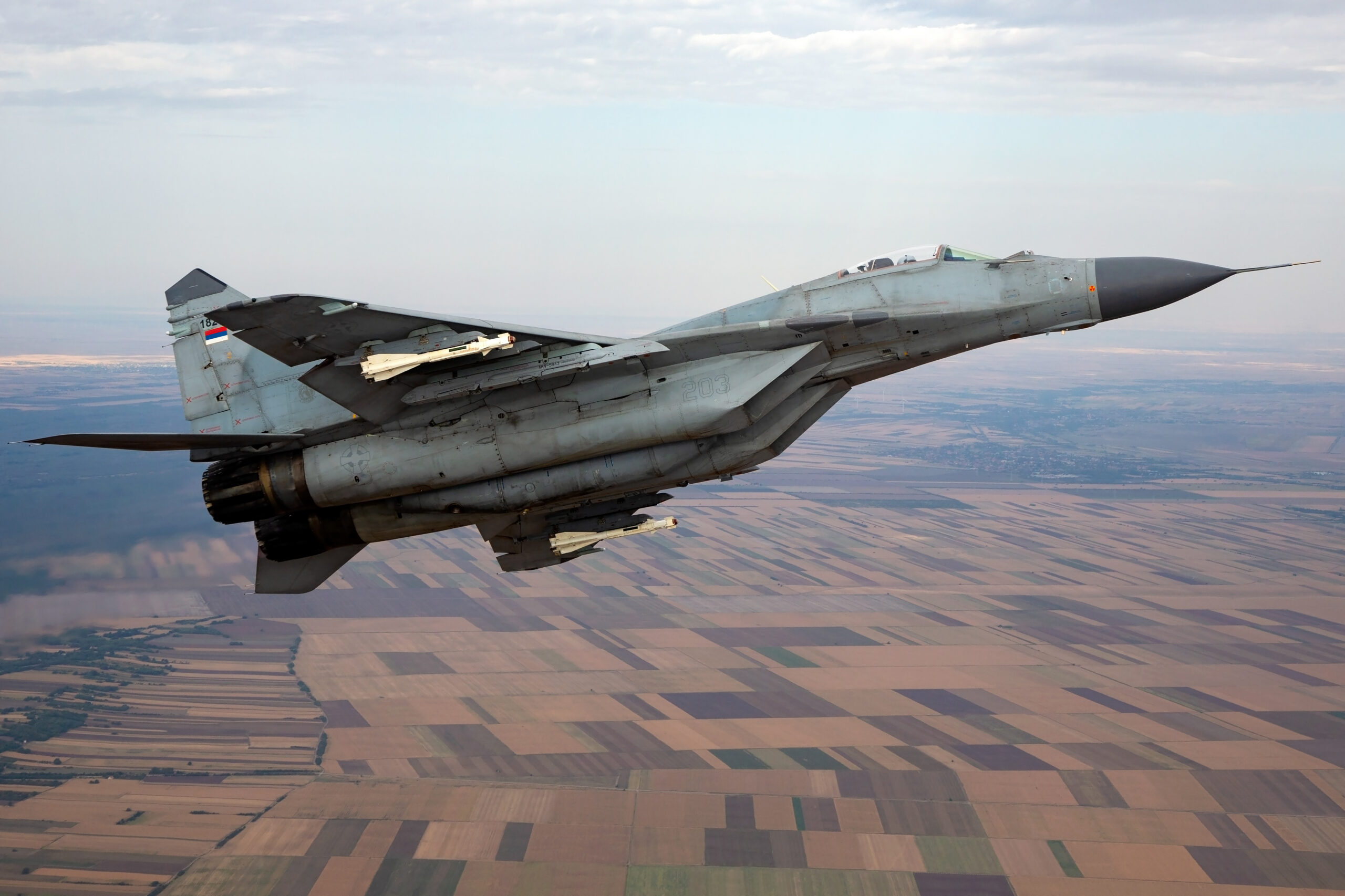 Serbian Air Force MiG-29 banking away over patchwork fields, missiles visible under the wing