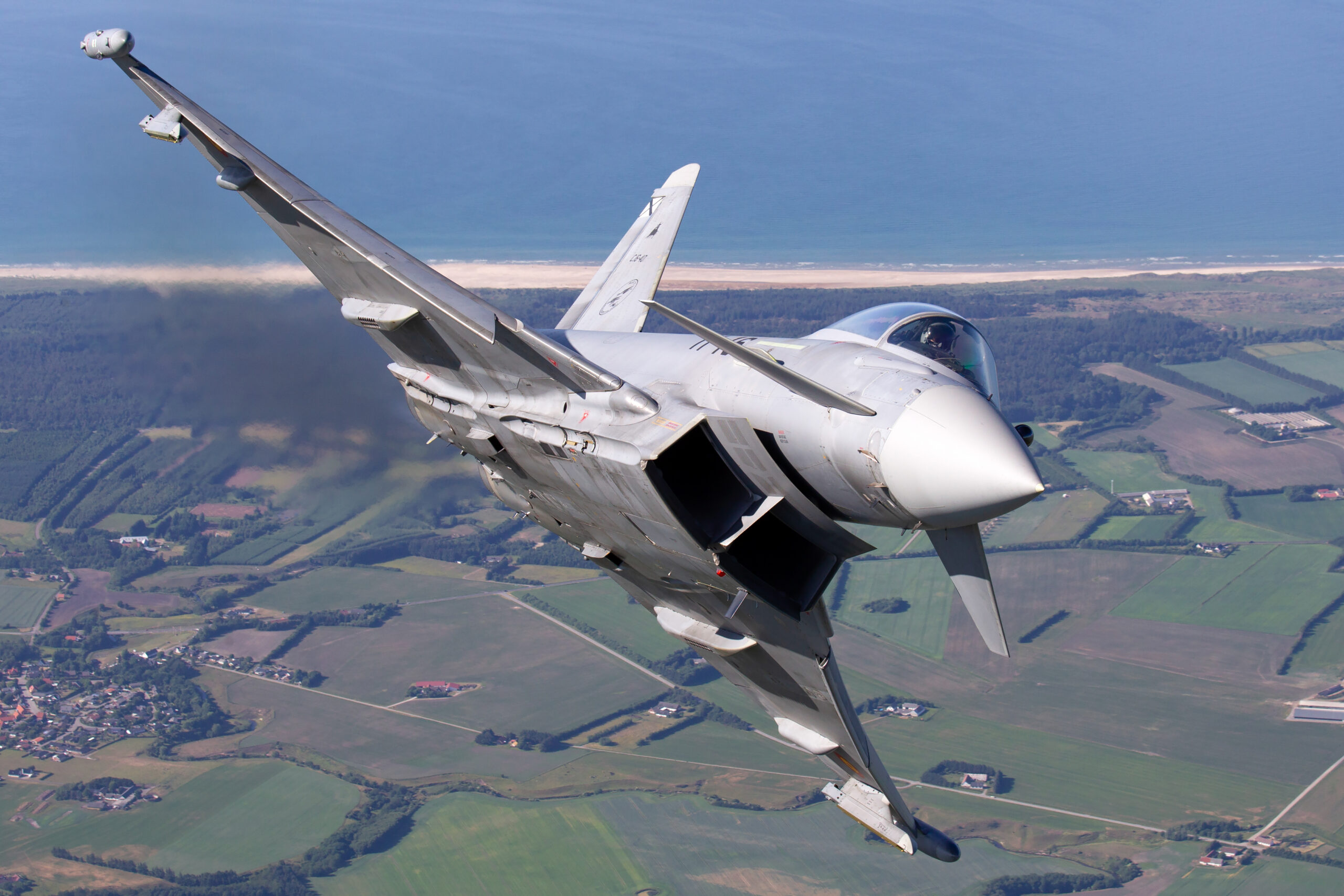 Spanish Air Force Eurofighter Typhoon 11-16 banking hard towards the camera over the Danish coastline, pilot visible and dark exhaust trailing