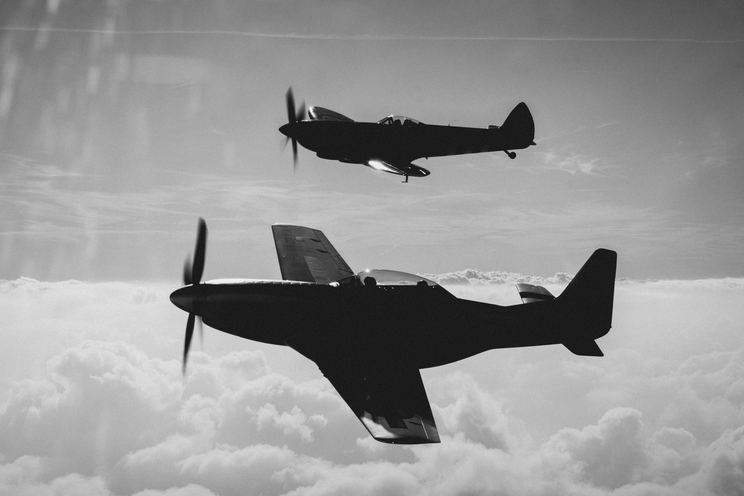 Black and white silhouette of a Spitfire and P-51 Mustang in formation above cloud