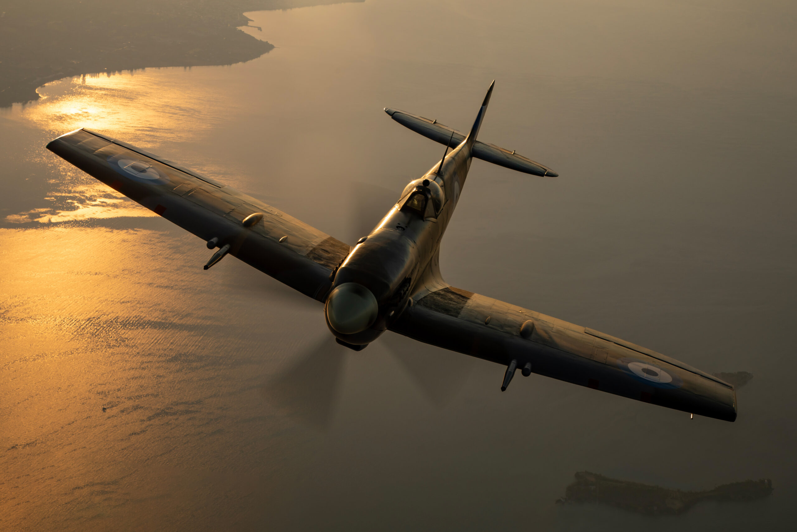 Spitfire banking in near-silhouette over a gold-lit coastline