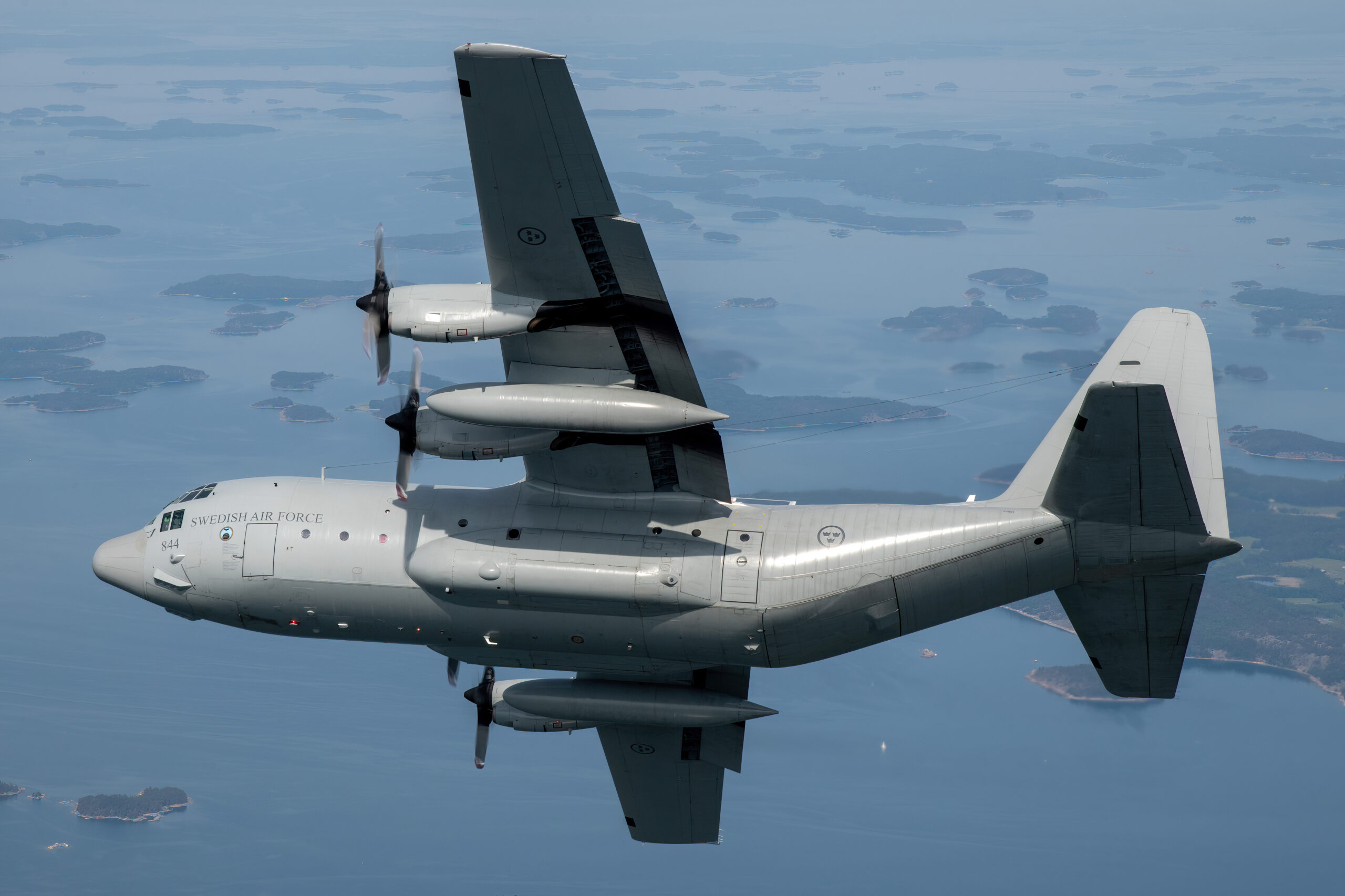 Swedish Air Force C-130H Hercules 844 banking over an island-dotted archipelago, all four propellers blurred into discs