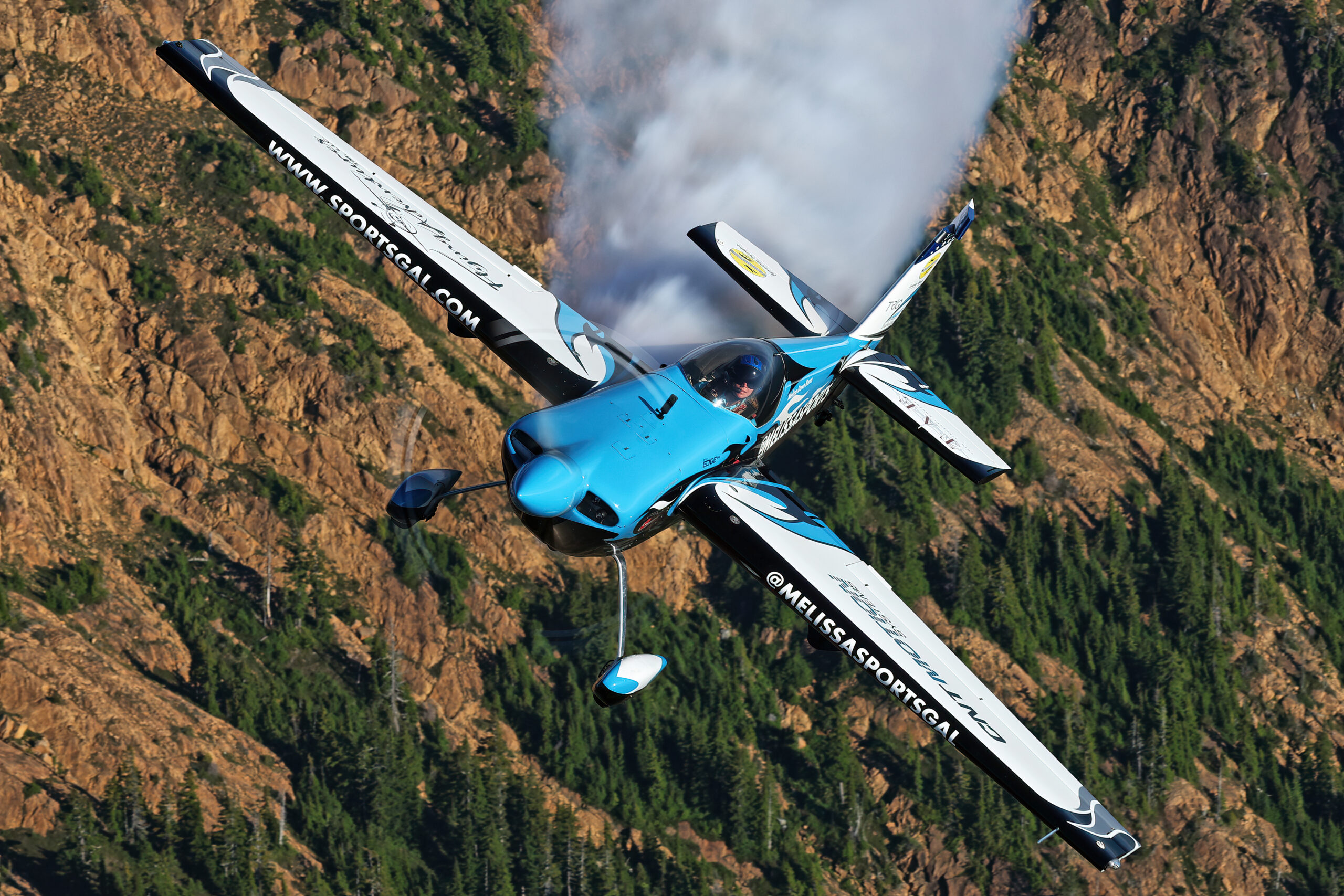 Blue and white Zivko Edge 540 aerobatic aircraft trailing smoke, banking over a rocky mountainside