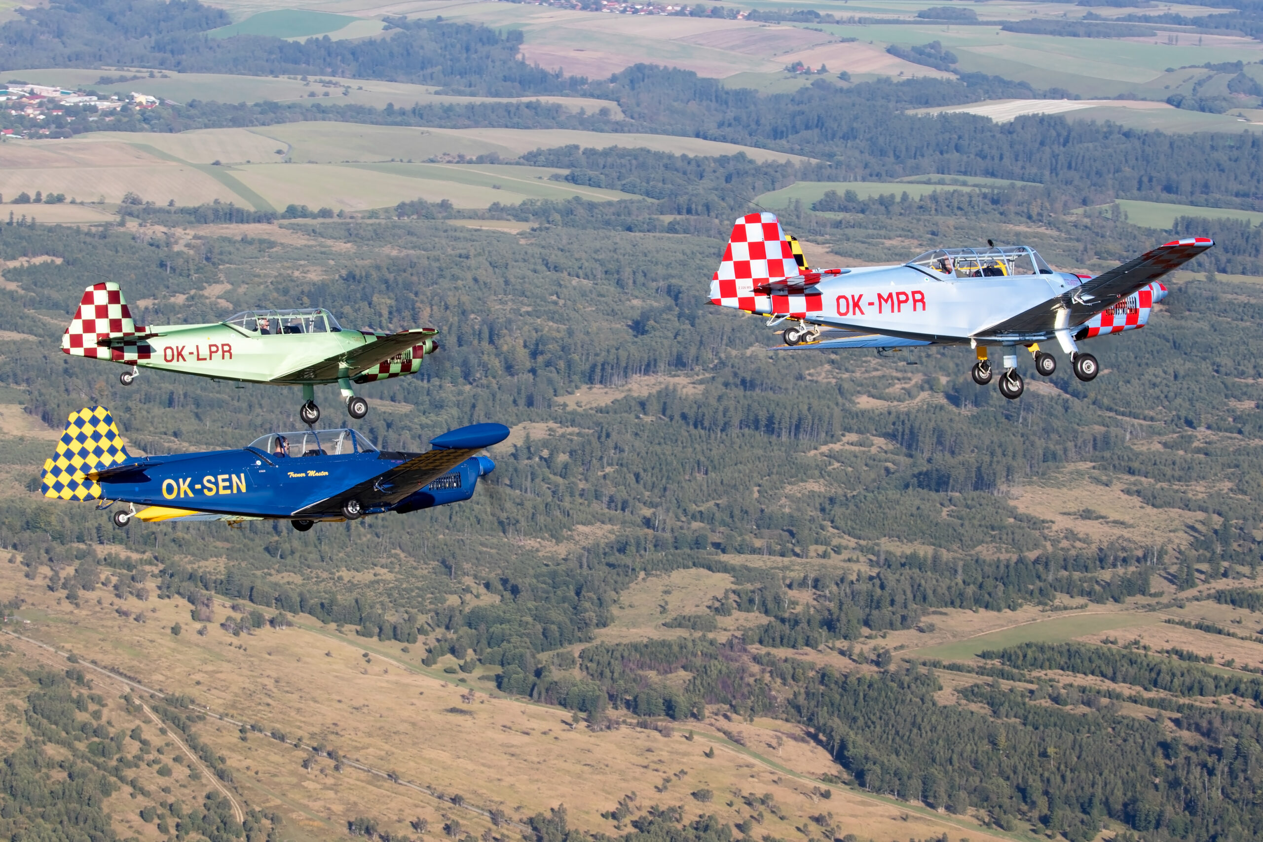 Three Zlin Treners in blue, green and grey checkerboard liveries, registrations OK-SEN, OK-LPR and OK-MPR, in formation over countryside