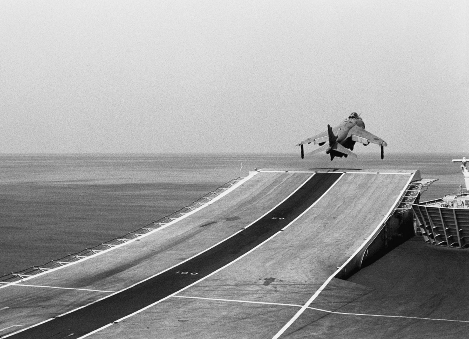 A Sea Harrier FRS1 lifting off the end of a carrier ski jump ramp, photographed from the deck looking forward over a grey sea