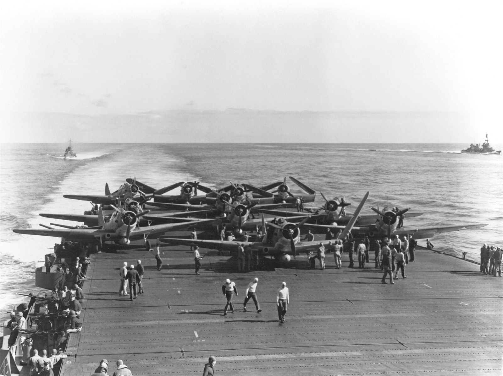 Eleven single-engined torpedo bombers with folded wings crowded on an aircraft carrier's wooden flight deck, deck crew standing among them, a cruiser visible on the horizon.
