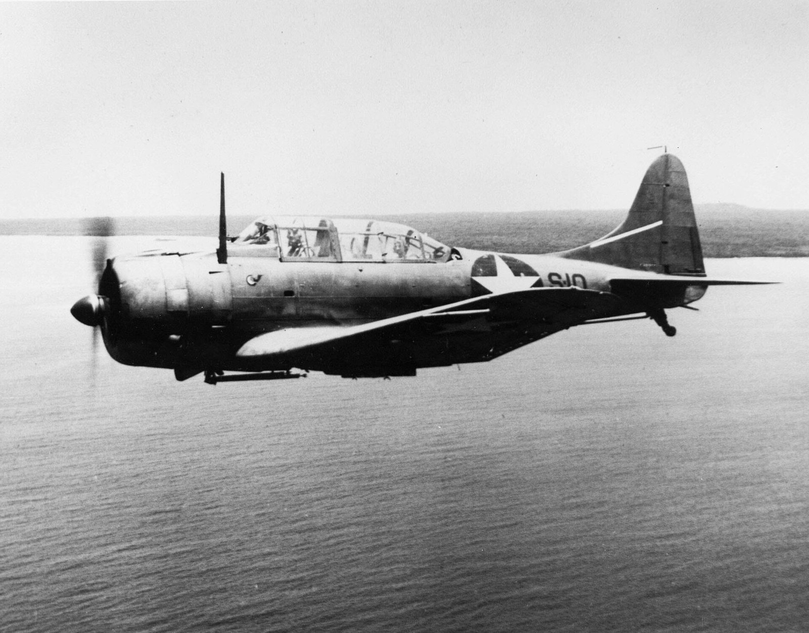A single-engined US Navy dive bomber in flight seen from above and behind, with a diagonal stripe on its rear fuselage and a squadron badge on the engine cowling.