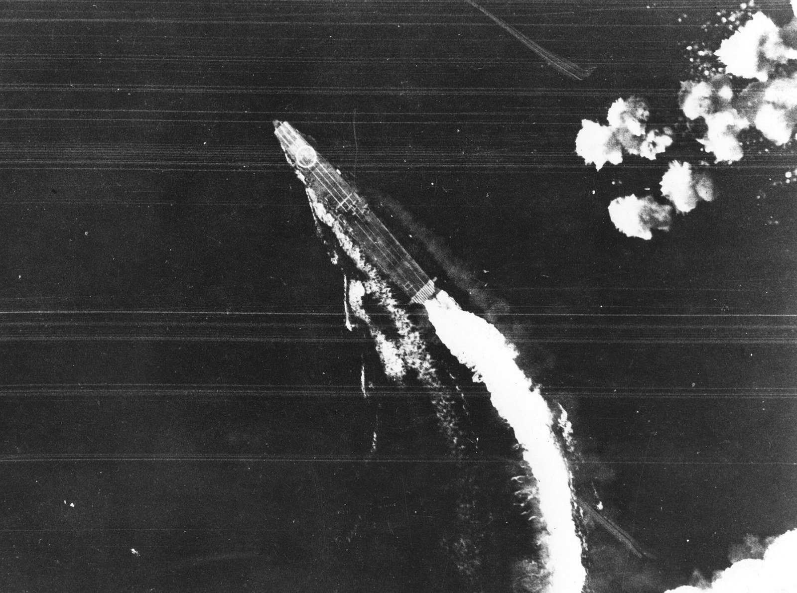 A Japanese aircraft carrier photographed from high above, turning hard to port and leaving a curved wake, with tall bomb splashes in the sea alongside her.