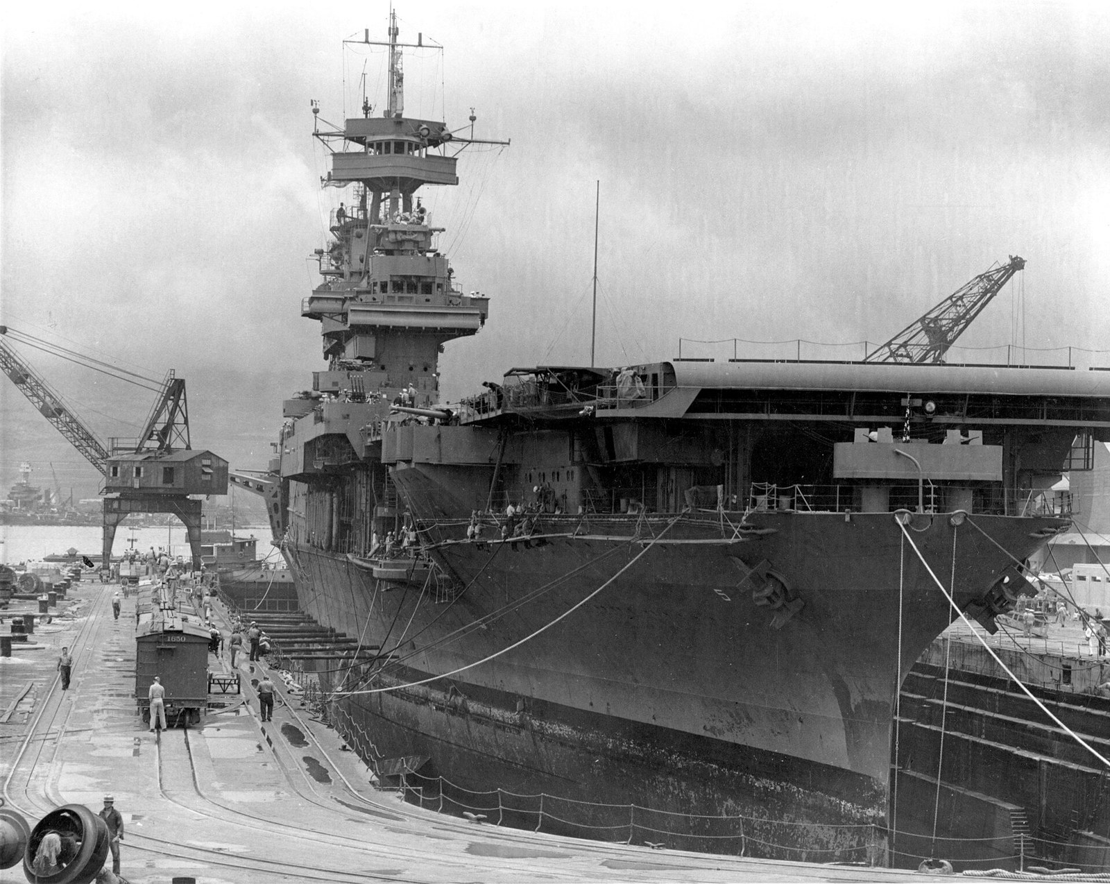 An aircraft carrier sitting high in a stone dry dock, her hull exposed to the keel blocks, with shipyard cranes alongside and a salvage operation on a sunken battleship in the distance.