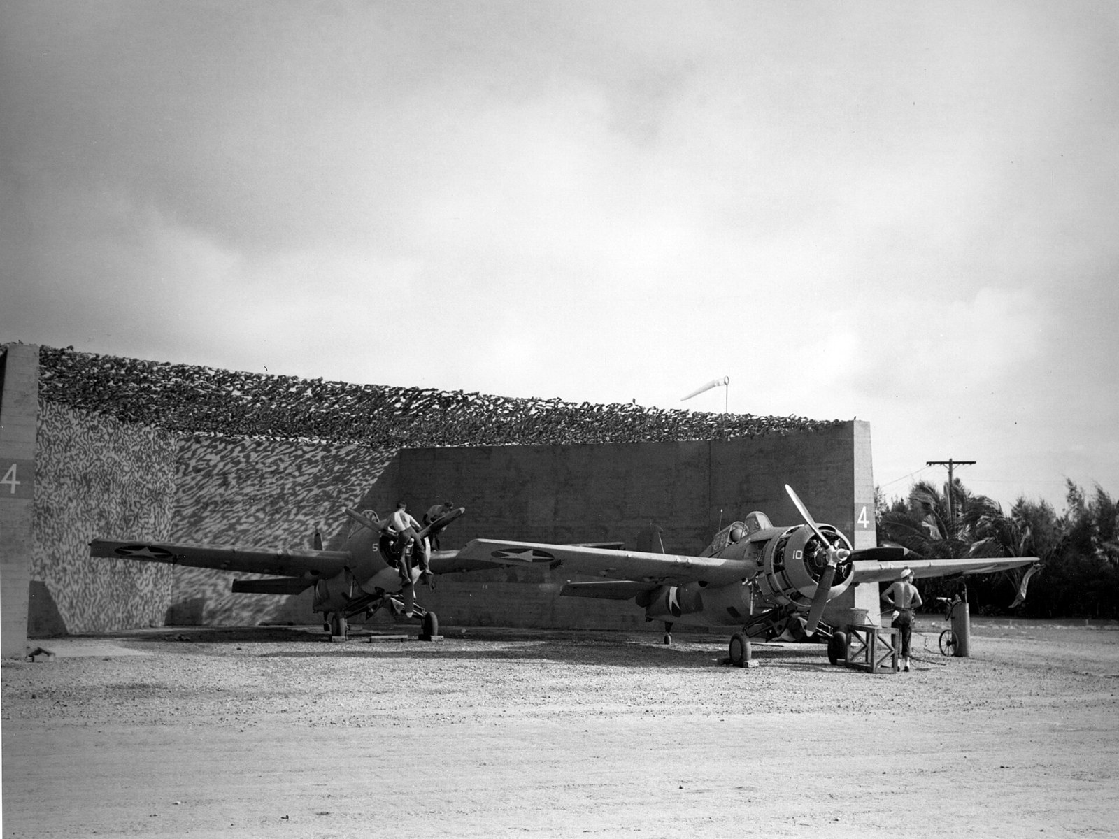 Two stubby US Navy fighter aircraft parked in an earth-banked revetment, ground crew working on the nearer aircraft's engine, camouflage netting overhead.