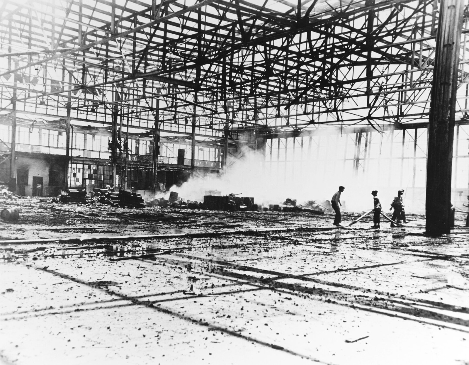 The gutted steel frame of a large hangar, its cladding burned away, with firefighters playing hoses on smoking wreckage and debris across the floor.