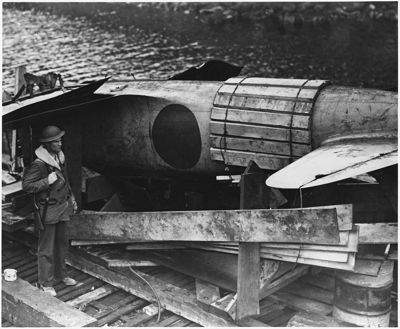 A Japanese single-seat fighter aircraft, its markings visible on the fuselage, resting on a barge under a tarpaulin sling in a coastal Alaskan setting.