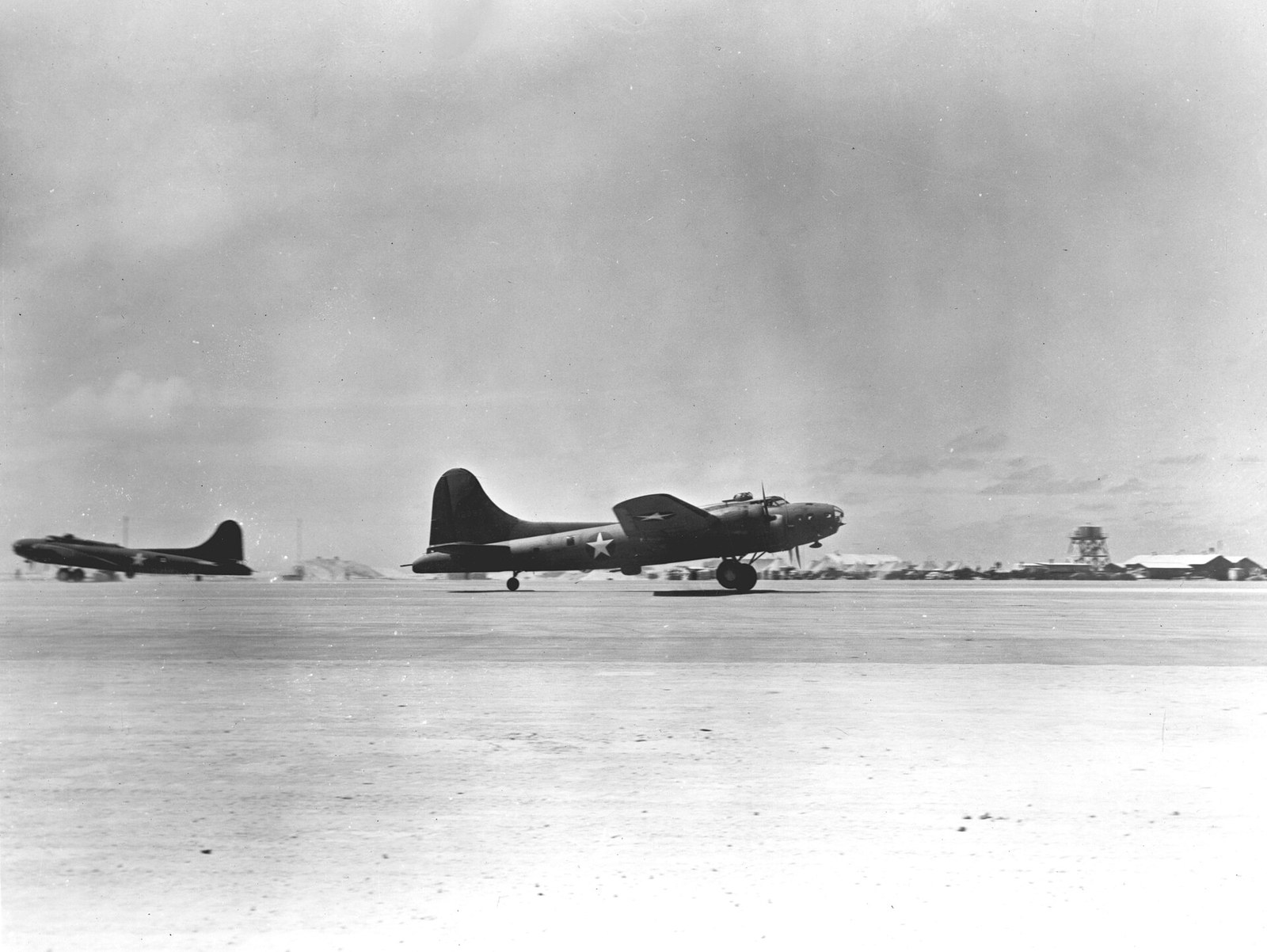 Four-engined US Army Air Forces heavy bombers lifting off from a low coral island airstrip, with flat scrub and the lagoon beyond.