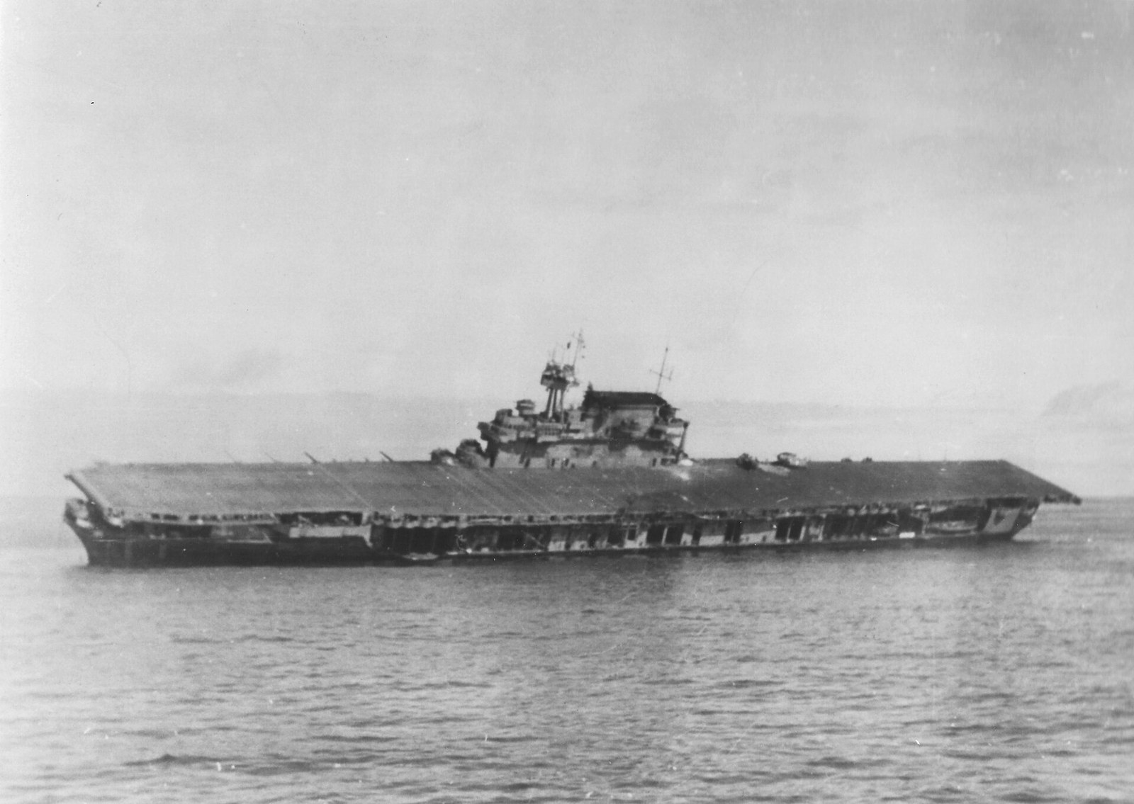 An aircraft carrier heeled far over to port and empty of her crew, seen from a nearby ship across open sea, two fighter aircraft still on her tilted flight deck.