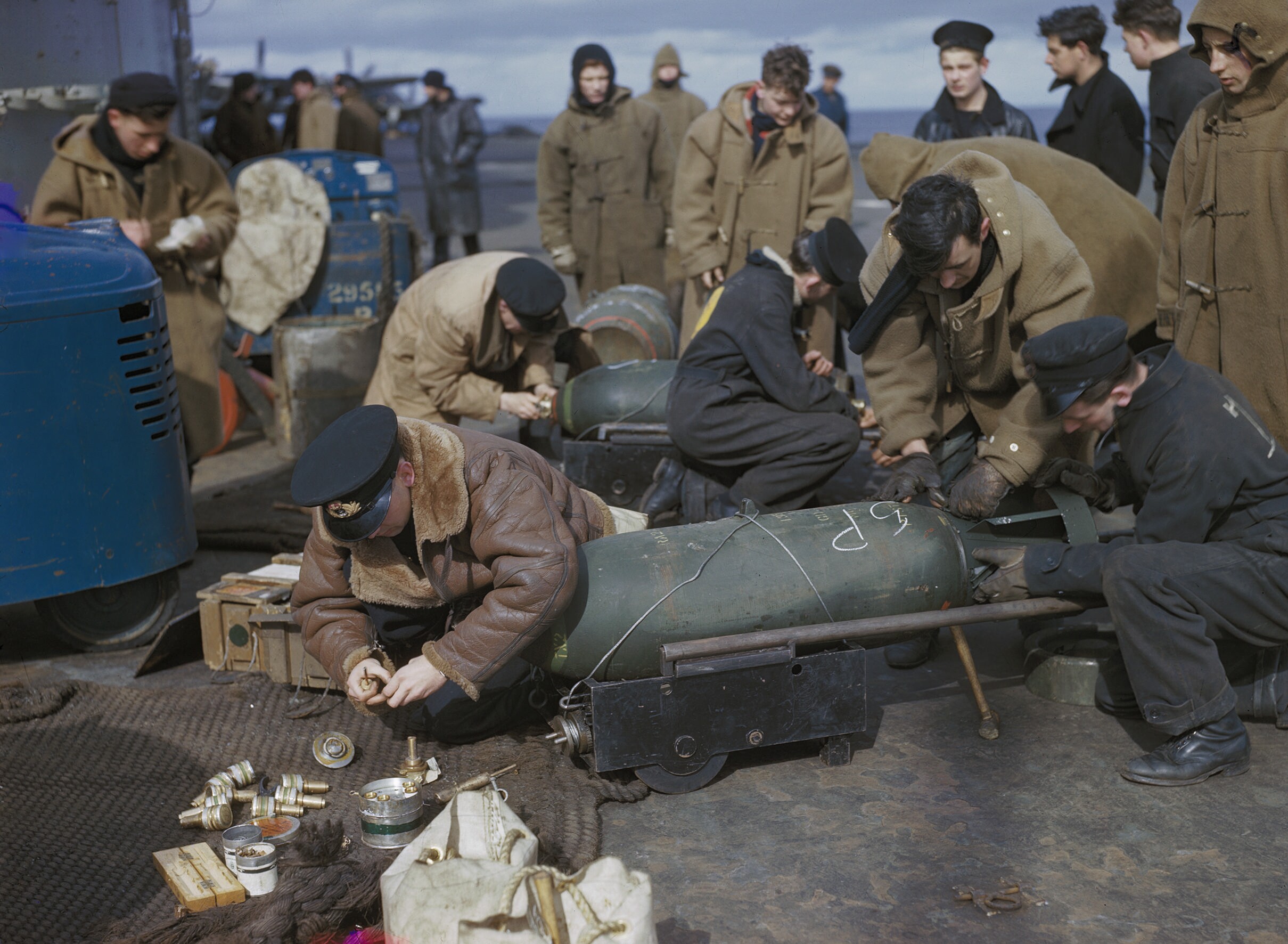 A colour photograph of Fleet Air Arm armourers in duffel coats fusing large armour piercing bombs on the flight deck of HMS Victorious
