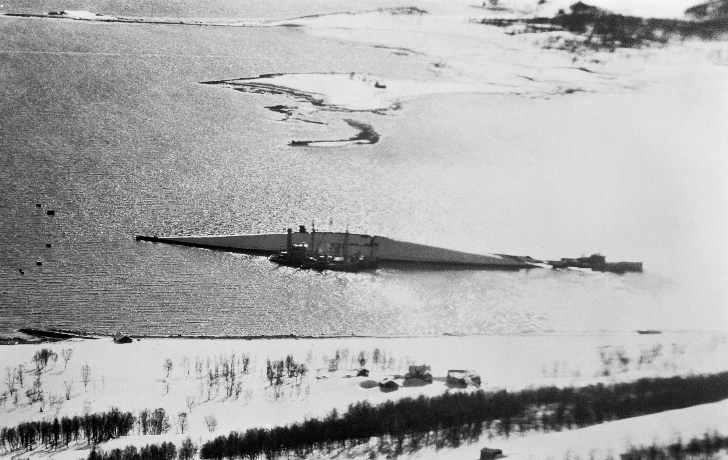 The capsized hull of the battleship Tirpitz lying bottom upwards in shallow water with a salvage vessel alongside