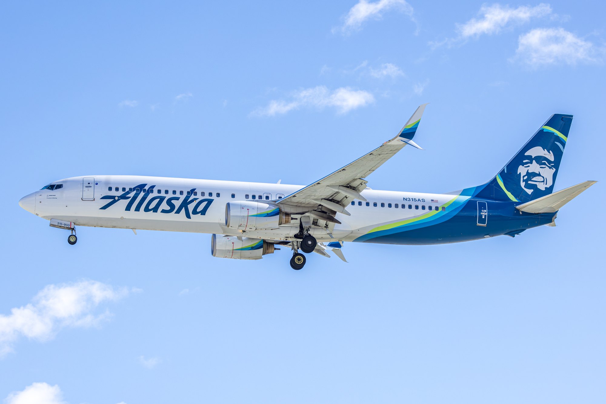 An Alaska Airlines Boeing 737-900, registration N315AS, gear down on final approach against a blue sky with light cloud.