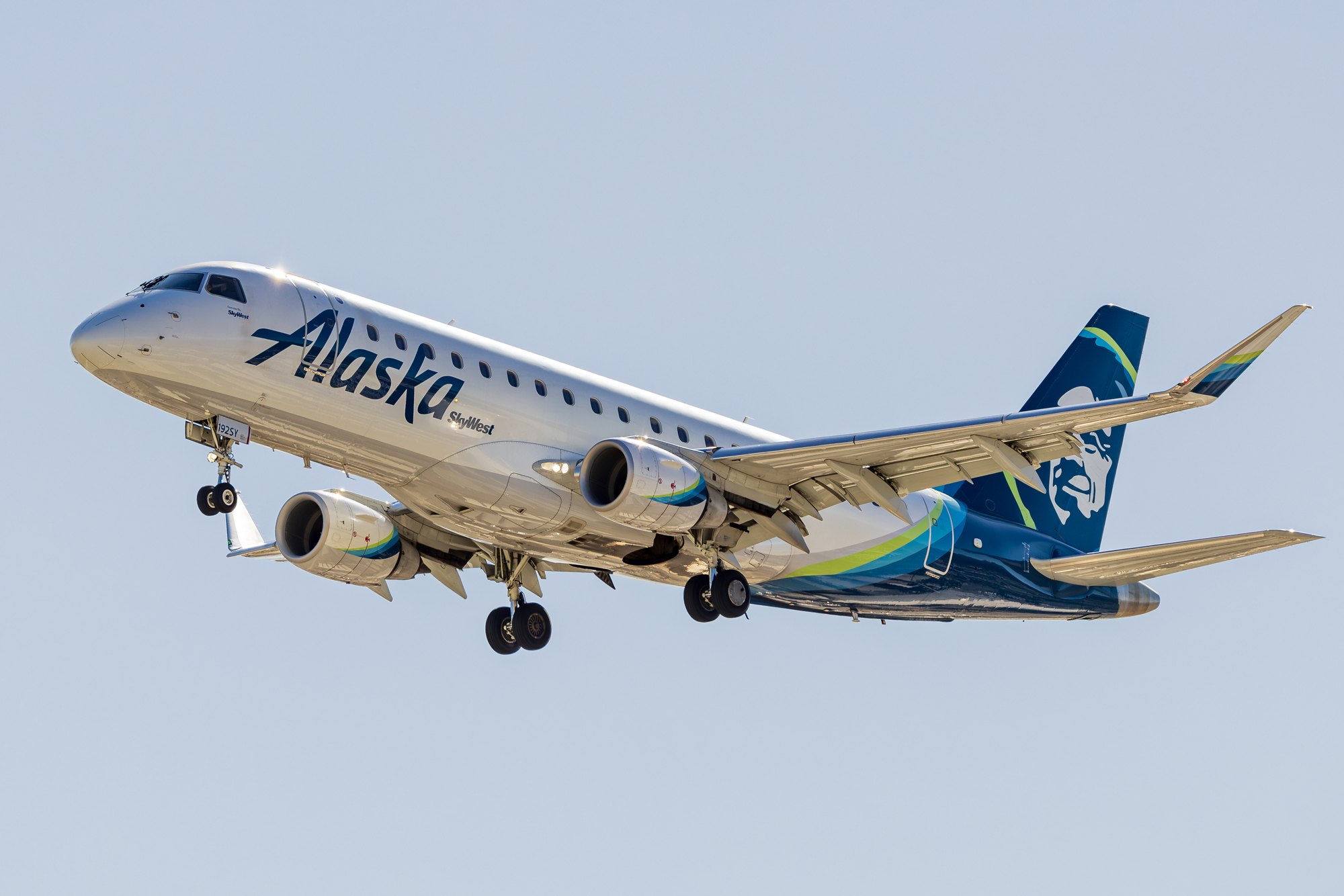 An Embraer E175 in Alaska Airlines colours with SkyWest titles, gear down on approach in warm low-angle light.