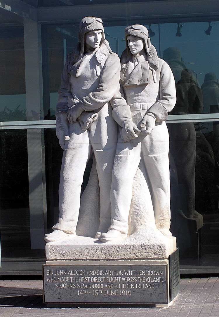 A stone statue of two airmen in flying kit standing side by side, on a plinth carved with the words who made the first direct flight across the Atlantic