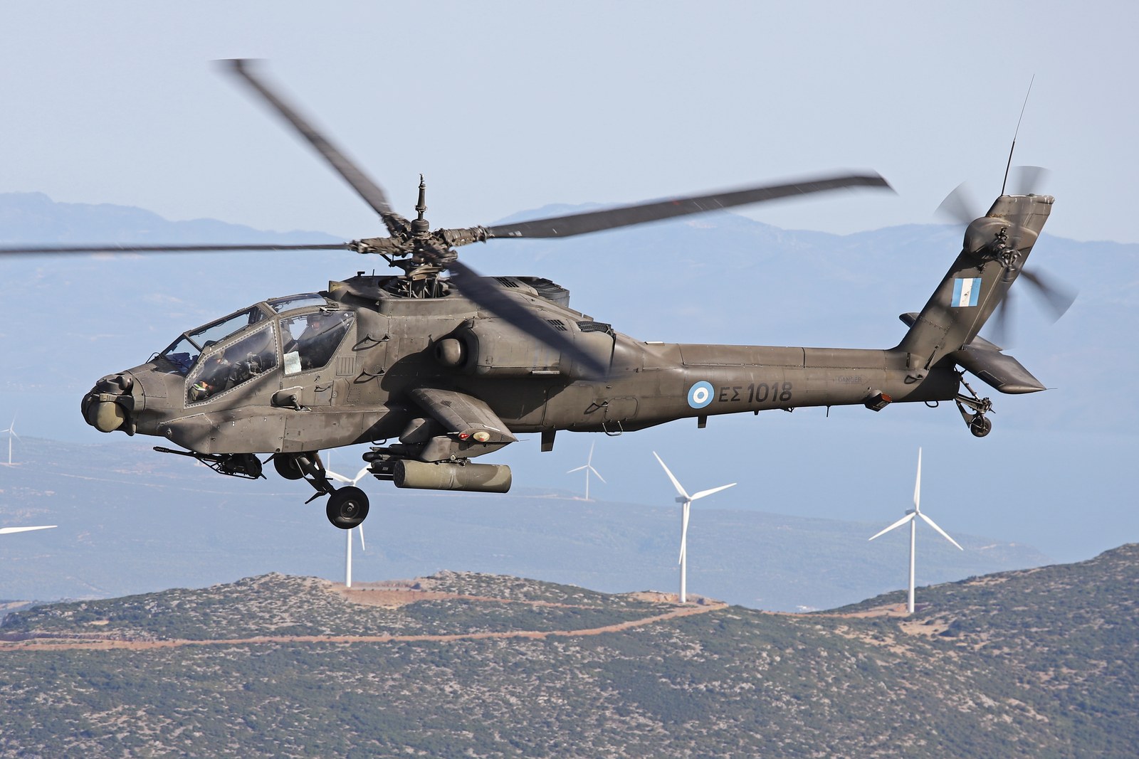 A McDonnell Douglas AH-64A Apache of the Hellenic Army in flight over Greek hills with wind turbines on the ridge behind it