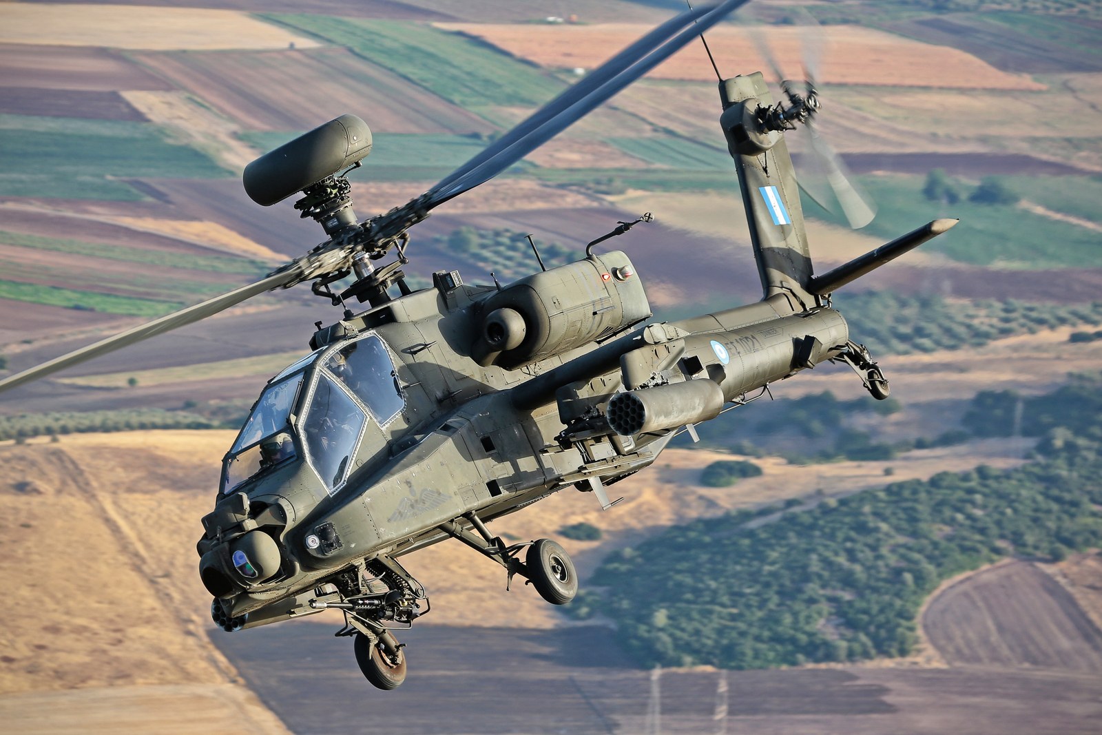 A Hellenic Army AH-64DHA Apache Longbow banking hard over farmland, turning its rotor disc towards the camera