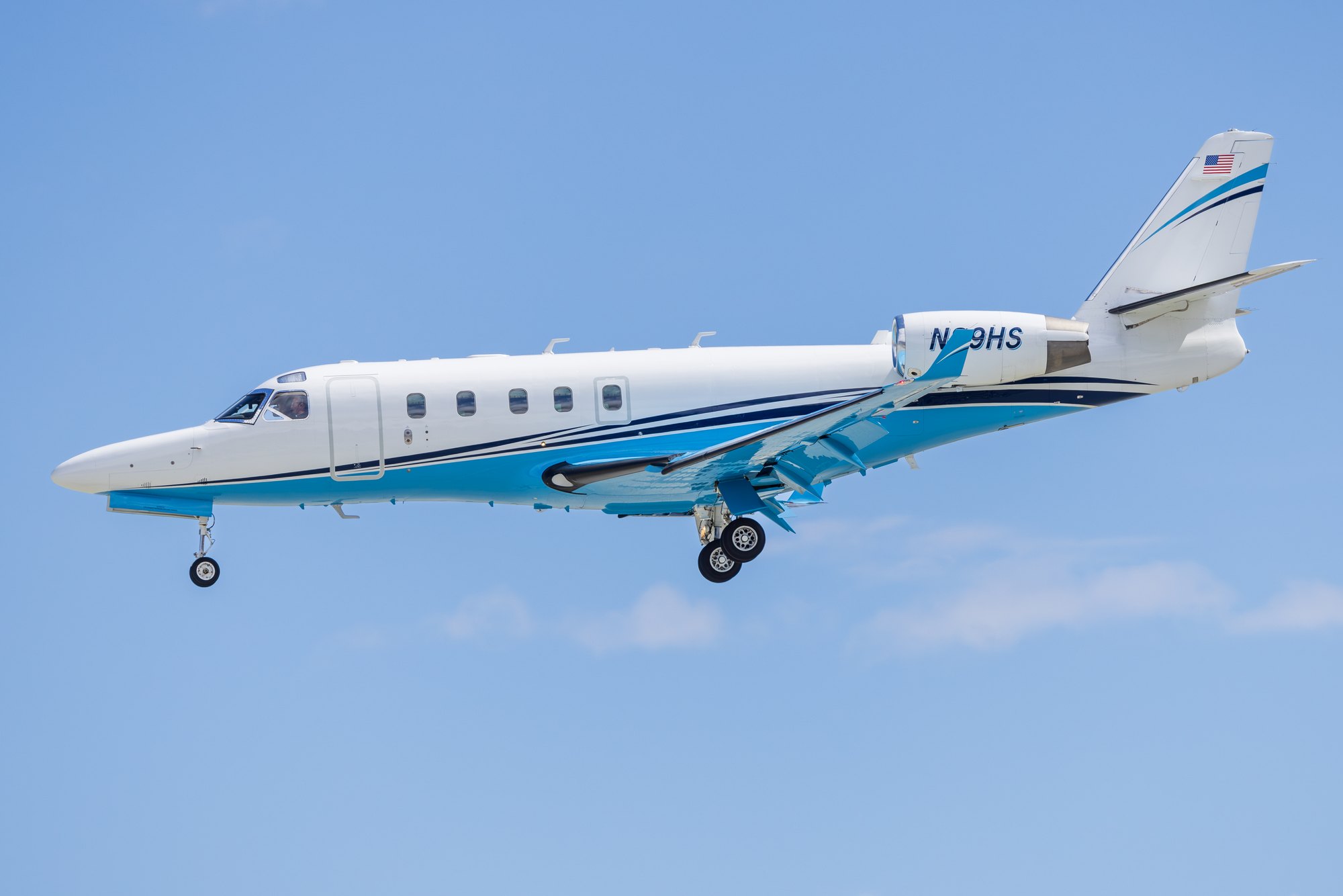 A white business jet with blue striping on approach at Palm Springs, gear down against a blue sky.