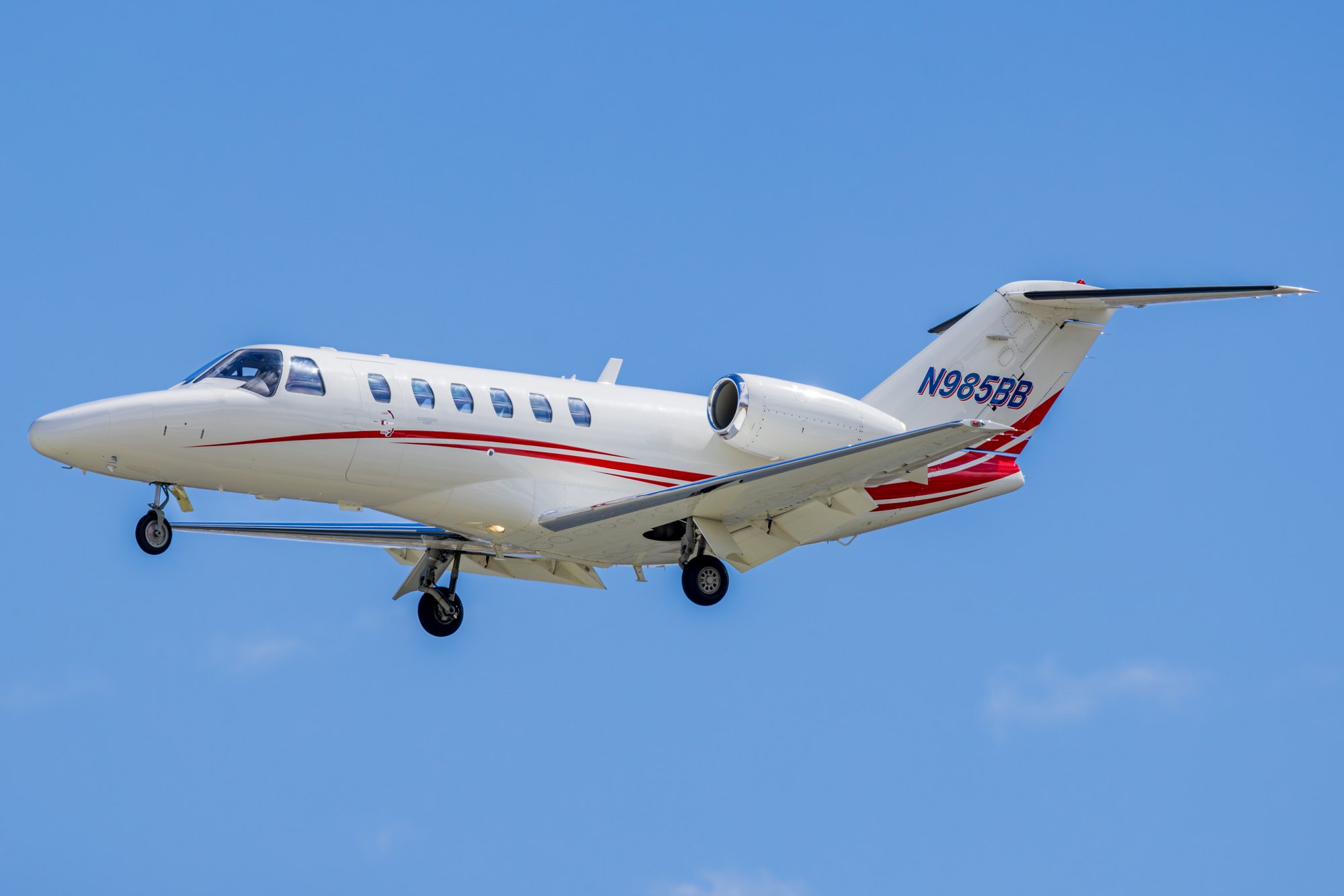 A white Cessna Citation CJ2 business jet with red and blue striping, registration N985BB, on approach with gear down against a clear blue sky.