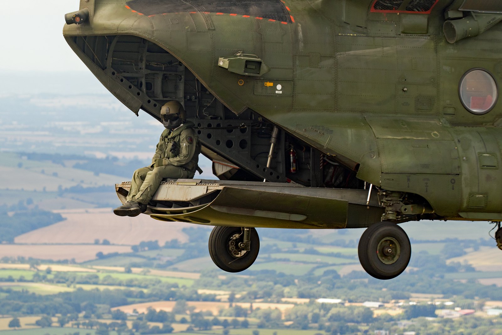 A helicopter crewman sitting on the edge of the open rear ramp of a Chinook in flight, with farmland far below