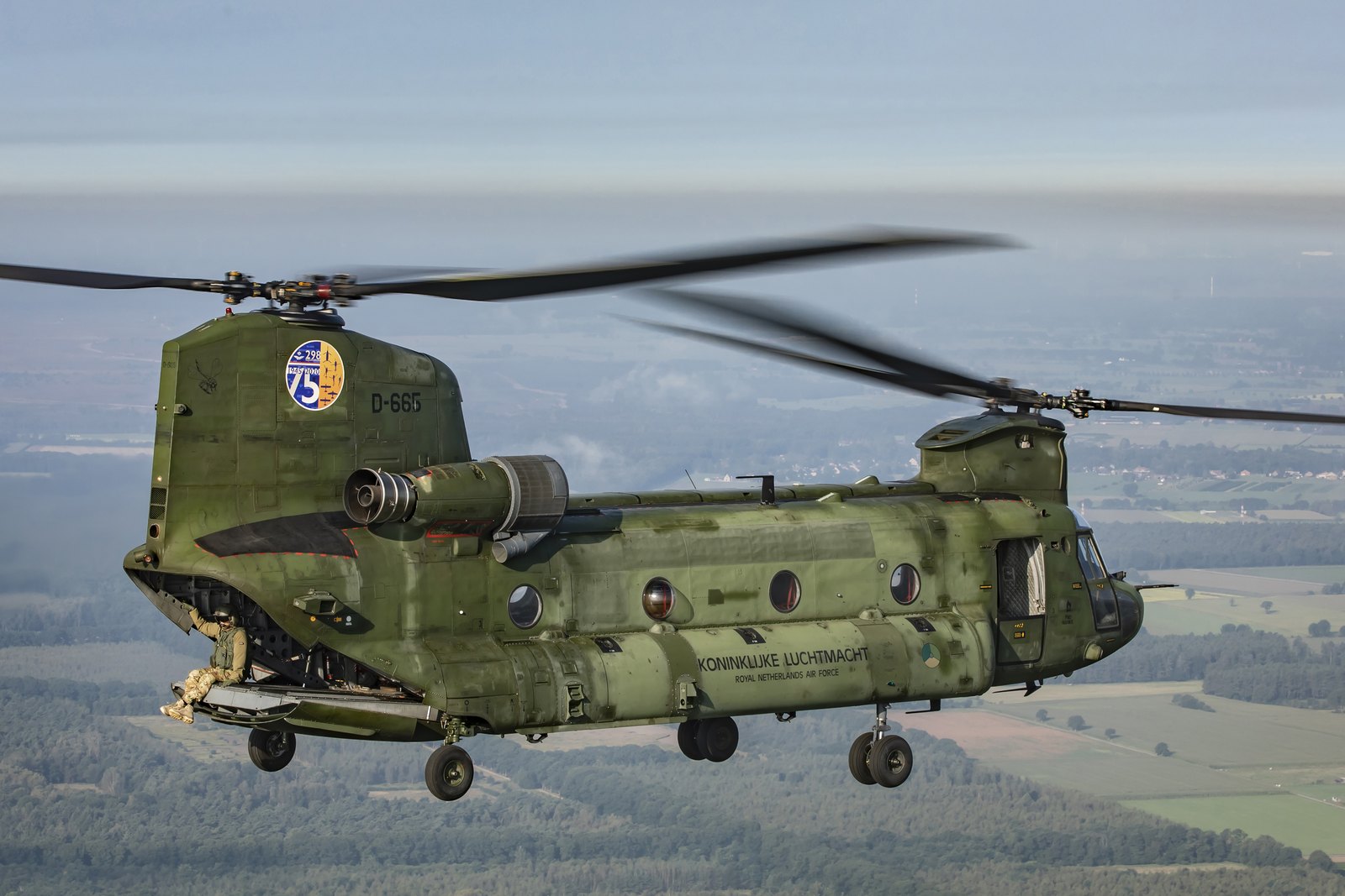 A crewman lying prone on the lowered rear ramp of a Royal Netherlands Air Force Chinook in flight