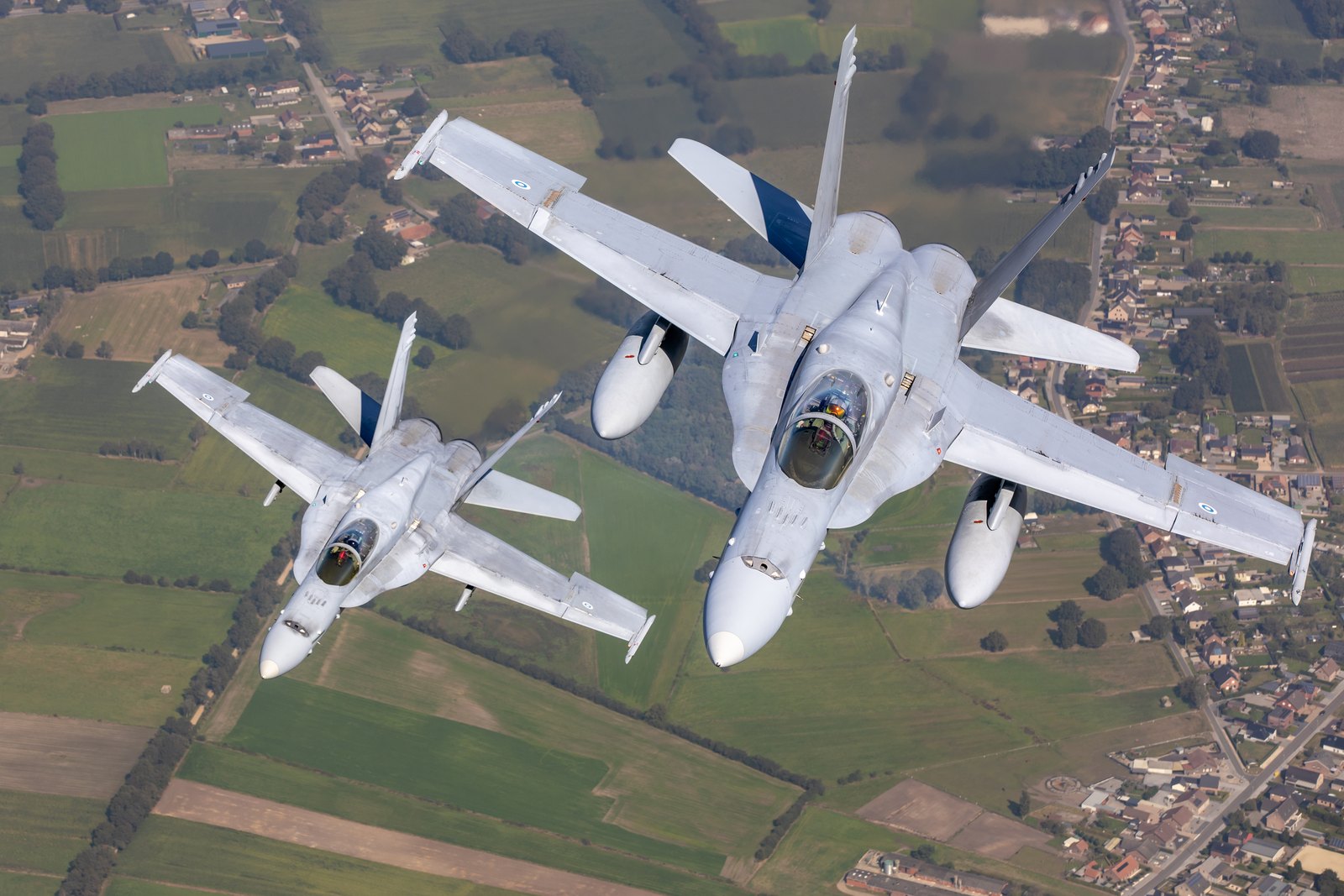 Two Finnish Air Force F/A-18C Hornets in echelon over Belgian farmland, each carrying a tank under either wing