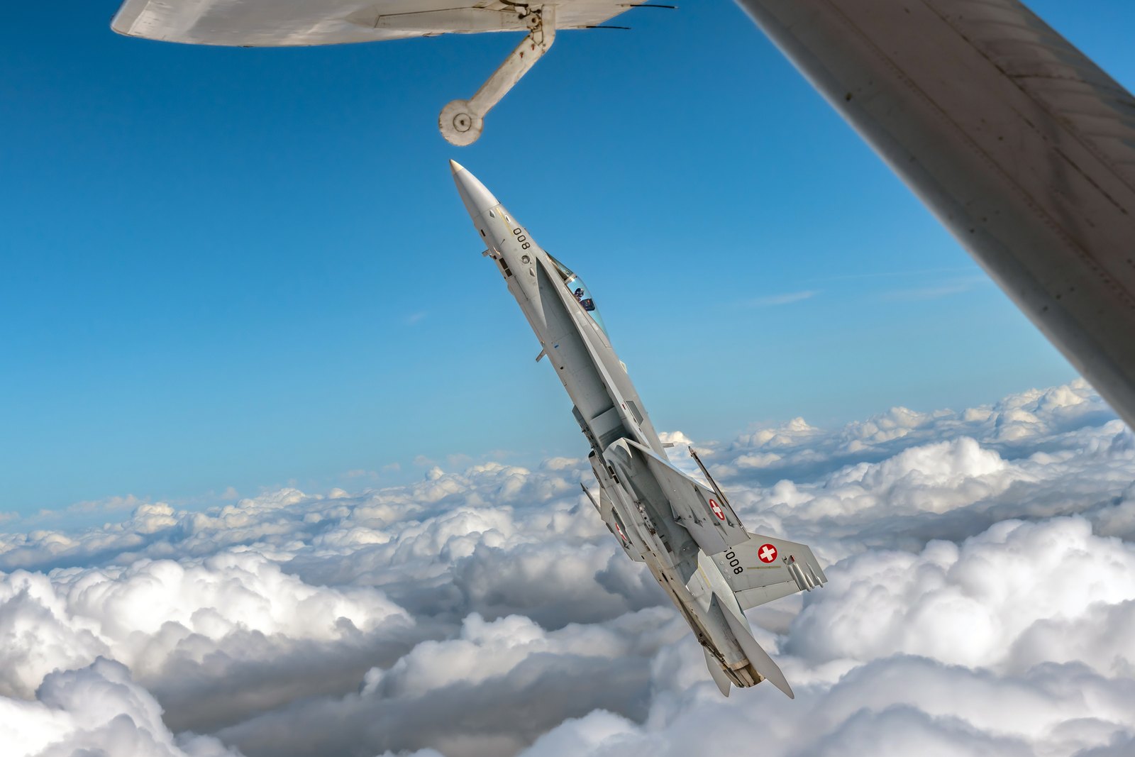 A Swiss Air Force F/A-18C Hornet climbing steeply above a cloud deck, seen past the wing of the camera aircraft