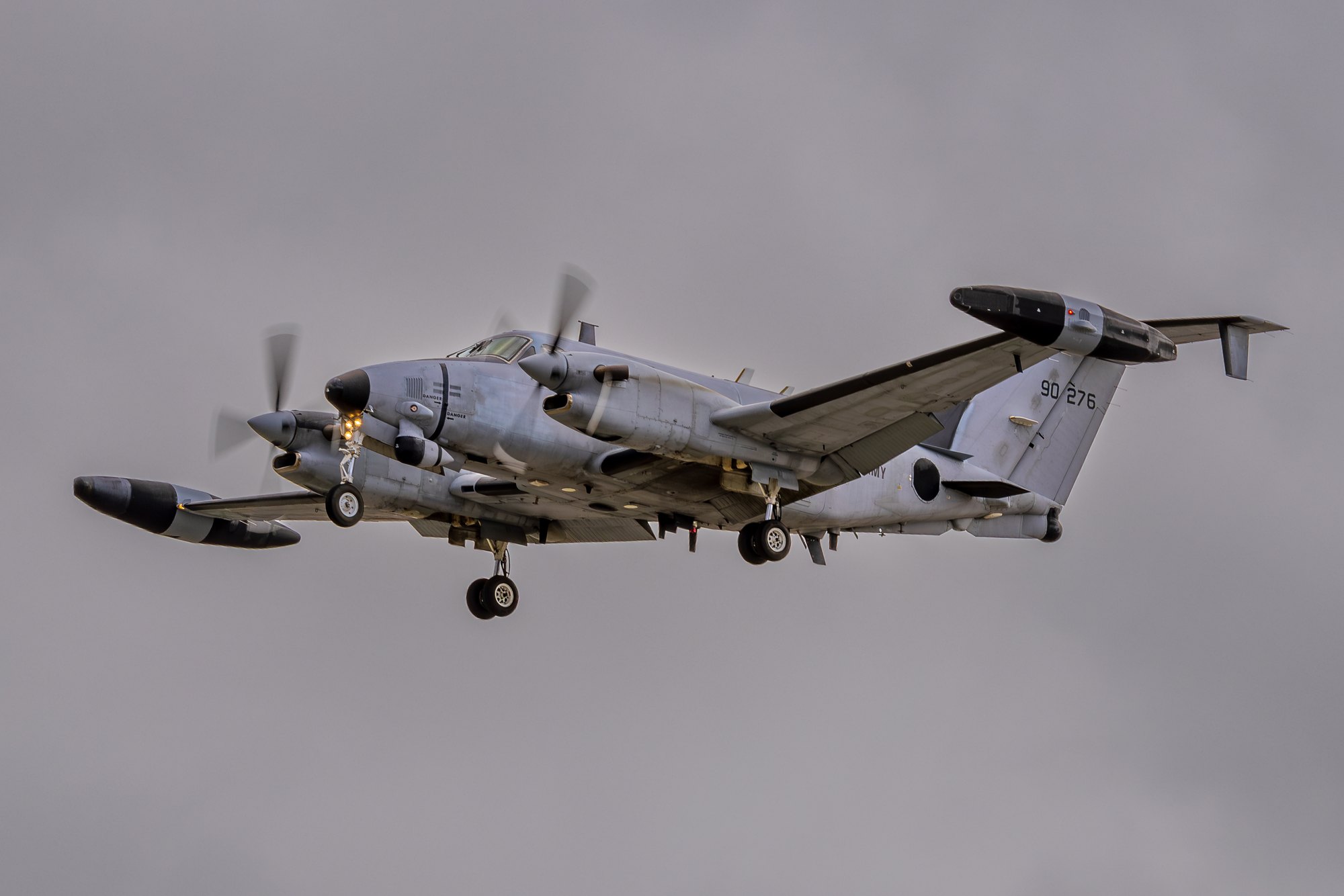 A grey US Army Beechcraft RC-12 Guardrail signals intelligence aircraft, serial 90-276, on approach with propellers blurred and wingtip pods and antennas visible.