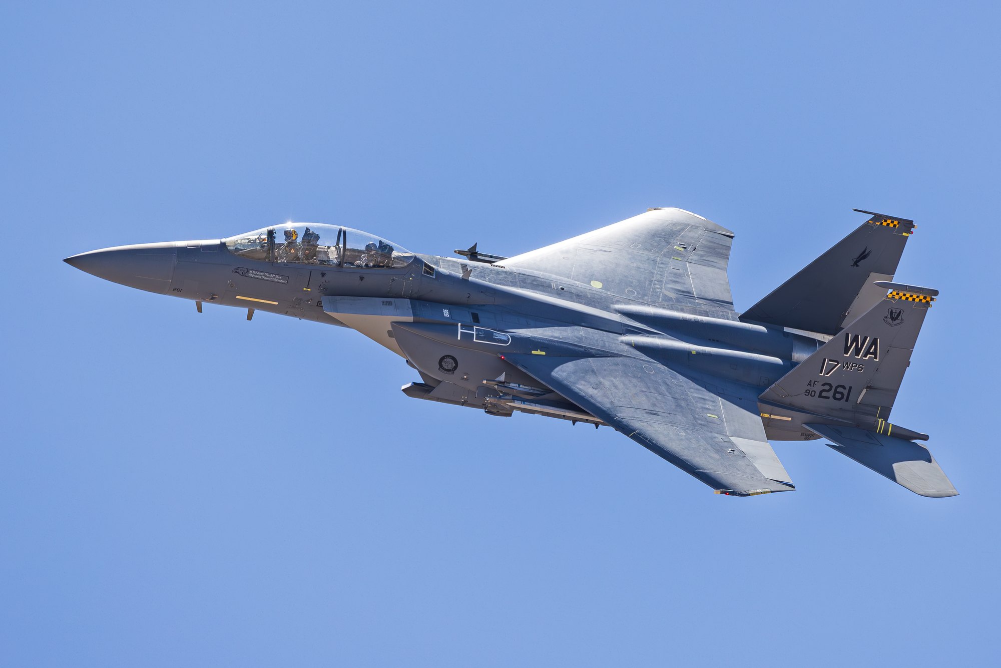 A US Air Force F-15E Strike Eagle, tail code WA with a yellow and black checked flash, serial 90-261, seen from above in a banking topside pass.
